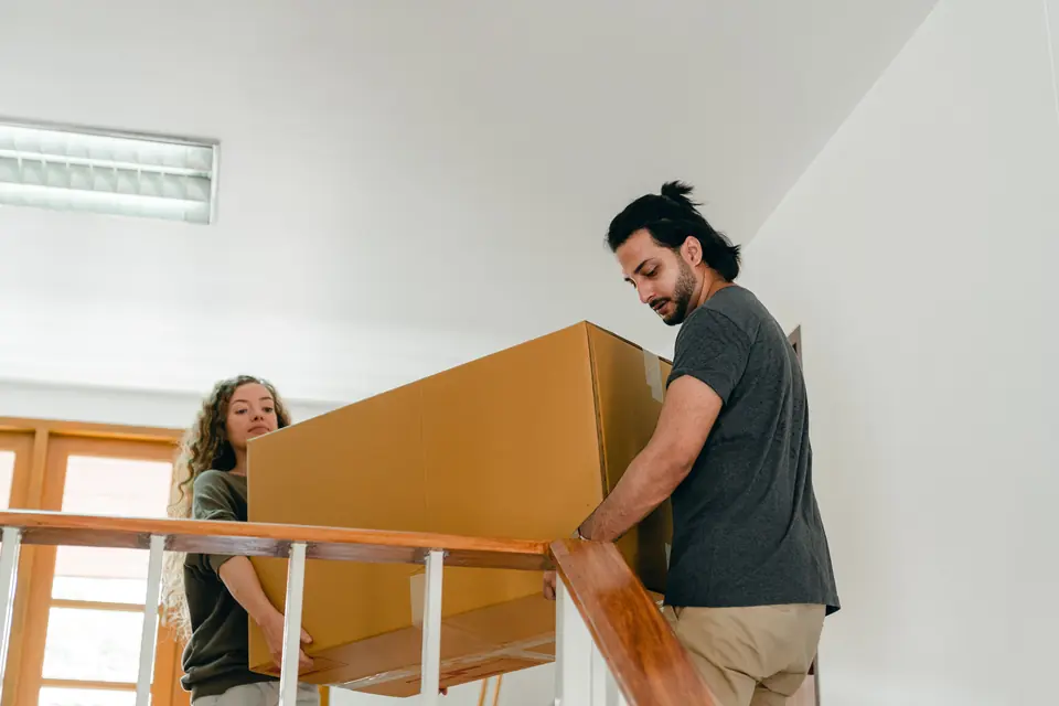 Zwei Personen tragen einen großen Umzugskarton die Treppe hinunter